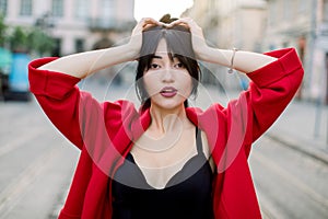 Outdoor portrait of young attractive confident Asian woman, wearing black top and stylish red blazer, posing on the