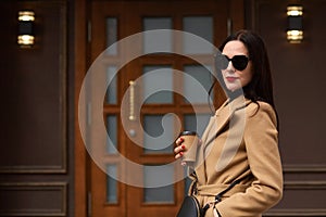 Outdoor portrait of a young attractive stylish woman wearing suitable accessories, beige trendy coat, holding a paper cup of