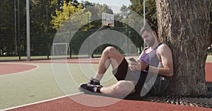Outdoor portrait of young bearded sportsman using cellphone at outdoor basketball court