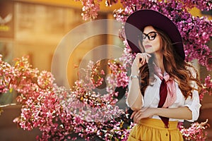 Outdoor portrait of young beautiful fashionable lady posing near flowering tree. Model wearing stylish accessories and