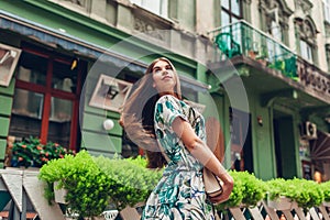 Outdoor portrait of young beautiful fashionable woman holding handbag and turning around by cafe. Girl walking in city