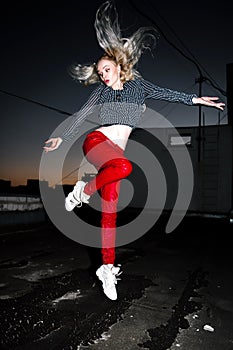 Outdoor portrait of young beautiful happy blond european lady posing on street at night. Model wearing stylish clothes red pants a