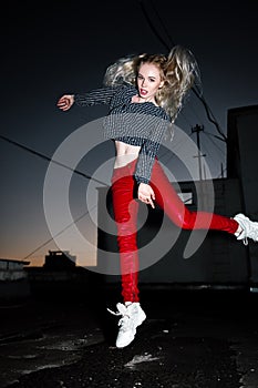 Outdoor portrait of young beautiful happy blond european lady posing on street at night. Model wearing stylish clothes red pants a