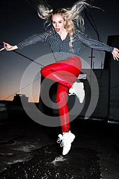 Outdoor portrait of young beautiful happy blond european lady posing on street at night. Model wearing stylish clothes red pants a