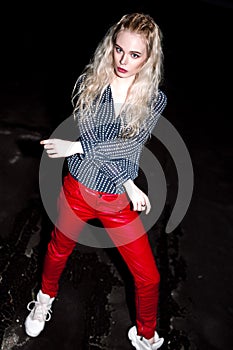 Outdoor portrait of young beautiful happy blond european lady posing on street at night. Model wearing stylish clothes red pants a