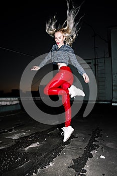 Outdoor portrait of young beautiful happy blond european lady posing on street at night. Model wearing stylish clothes red pants a