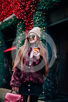 Outdoor portrait of young beautiful happy smiling girl posing on street. Model wearing stylish warm clothes.Keeps coffee. concept