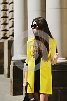 Outdoor portrait of young beautiful lady walking on the street. Model wearing sunglasses and stylish yellow summer dress
