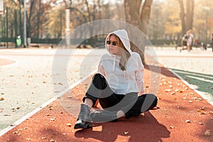 Outdoor portrait of young beautiful woman with long in sunglasses and a white hooded sweater sitting on the sportsground track