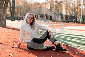 Outdoor portrait of young beautiful woman with long in sunglasses and a white hooded sweater sitting on the sportsground track