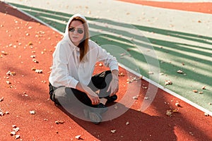 Outdoor portrait of young beautiful woman with long in sunglasses and a white hooded sweater sitting on the sportsground track