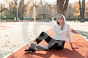 Outdoor portrait of young beautiful woman with long in sunglasses and a white hooded sweater sitting on the sportsground track