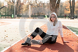 Outdoor portrait of young beautiful woman with long in sunglasses and a white hooded sweater sitting on the sportsground track