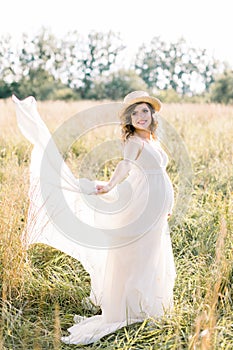 Outdoor portrait of young pregnant woman in the field. Pretty pregnant woman in white dress and hat relaxing in the