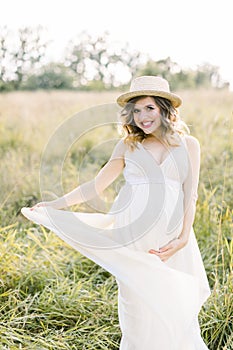 Outdoor portrait of young pregnant woman in the field. Pretty pregnant woman in white dress and hat relaxing in the