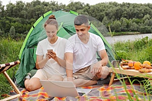 Outdoor portrait of young pretty hiker couple sitting near a tent looking at laptop in the nature, woman using cell phone, family