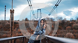 Outdoor portrait of young, thoughtful hooded girl, standing on old rusty bridge in rainy day.