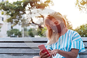 Outdoor portrait of a young woman with a protective mask and cell phone. Girl with a cell phone sitting in the park