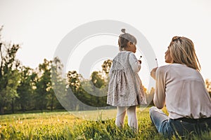 Outdoor rear view of happy girl kid playing with her mother outside. Portrait of woman and her cute child blowing dandelion in the