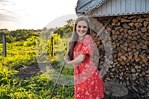 Outdoor right sunnny portrait of happy teen red head girl in red dress that standing with rakes and looking at the