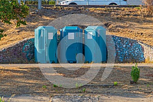 Outdoor scene with three blue porta-potties in a triangular formation, against a rocky backdrop suggesting a rugged photo