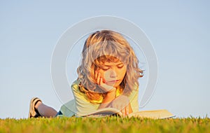 Outdoor school, motivation learning kids. Child boy reading book, laying on grass in field on sky background. Portrait