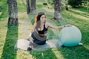 Outdoor shot of active sporty woman takes break after physical exercises, poses on fitness mat with fitball, enjoys fresh air in