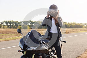 Outdoor shot of attractive confident moto driver with helmet wearing for safety, sitting on her motorbike at road, having stop,
