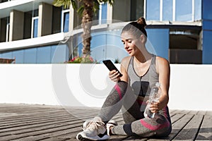 Outdoor shot of attractive fitness woman sitting on wooden pier with smartphone, having break after workout, drinking