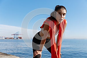Outdoor shot of attractive sportswoman having a break after running on the seaside promenade. Woman taking a breath and