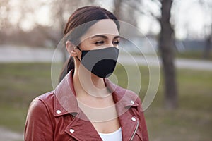 Outdoor shot of beautiful brunette woman wearing leather jacket and black protective mask, looking in distance, protecting herself