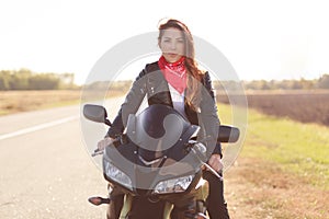Outdoor shot of beautiful motorcyclist has long journey on her motorbike, wears red stylish bandana and leather jacket, likes extr