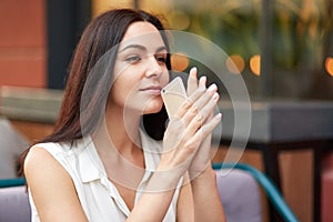 Outdoor shot of brunette female with serious expression, holds smart phone, waits for important call, being deep in thoughts, spen