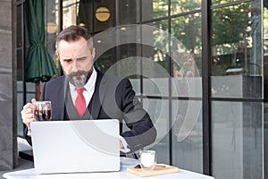 Outdoor shot of Caucasian businessman drinking hot morning coffee at the coffee shop while using computer laptop to work and check
