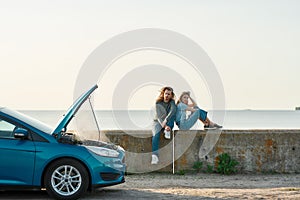 Outdoor shot of couple of travelers sitting near the broken car with open hood and smoking engine, man and woman looking