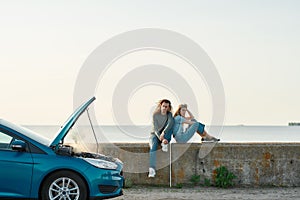 Outdoor shot of couple of travelers sitting near the broken car with open hood and smoking engine, man and woman looking