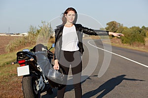 Outdoor shot of female biker hitchhikes on road, holds helmet in one hand, wears leather jacket, stops other transport, needs help