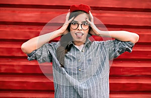 Outdoor shot of funny brunette young woman with hands on head, dressed in casual blue shirt, red beret hat and glasses, making