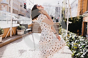 Outdoor shot of graceful girl relaxing in sunny day. Stylish short-haired lady in white dress dancing on the street..