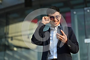 Outdoor shot of happy young businessman standing standing outdoors in urban modern city and using mobile phone