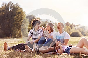 Outdoor shot of happy youngsters have charming smiles, look into distance, sit on plaid, pose against green nature and sunshine ba