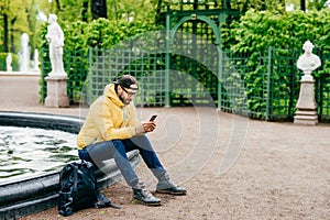 Outdoor shot of hipster guy with beard dressed stylish resting in park near fountain surfing social networks using his cell phone