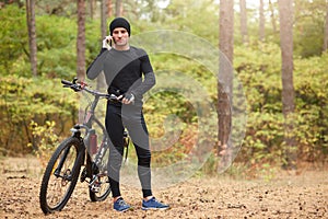 Outdoor shot of man wearing black track suit and sneakers, standing near his mountain bike and speaks phone with beautiful forest