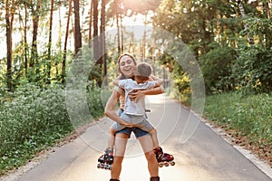 Outdoor shot of mother with little boy kid roller skating in summer park, mother holding child in hands, hugging and smiling with