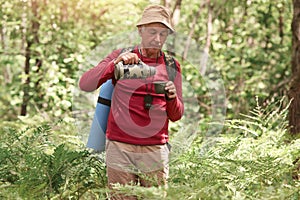 Outdoor shot of old man with bag on back, holding thermos and being ready to drik hot tea or coffee, having sleeping pad, wearing