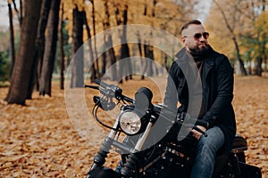 Outdoor shot of pensive male biker keeps gaze aside, has thoughtful look, wears sunglasses, sweater, black jacket and jeans, poses