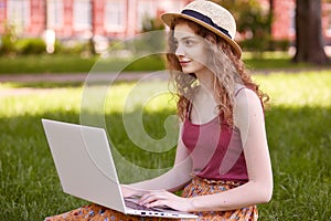 Outdoor shot of pretty young woman sitting in park with laptop on legs, spending summer day working outdoor, looking at distance,
