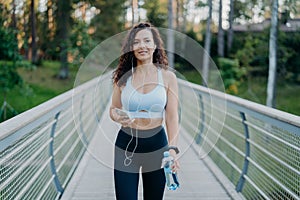 Outdoor shot of slim woman in active wear goes jogging near forest, enjoys listening music in earphones from favorite playlist,