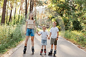 Outdoor shot of smiling attractive female with her little sons standing on road in summer park and holding hands, family