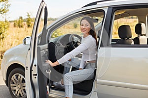 Outdoor shot of smiling satisfied woman with dark hair getting out of car with happy positive facial expression, female wearing